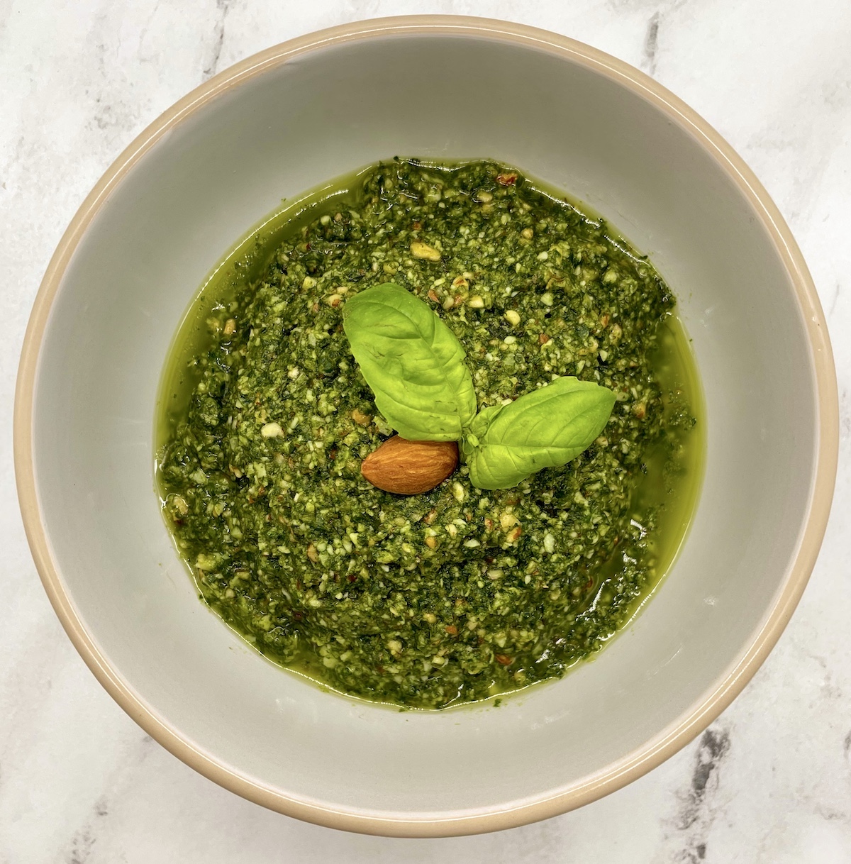 Bright green almond pesto in a white bowl on a white marble counter garnished with fresh basil leaves and an almond