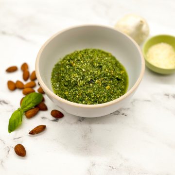 Bright green almond pesto in a white bowl on a white marble counter garnished with fresh basil leaves and an almond