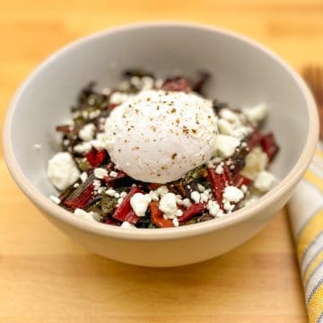 Brown rice grain bowl with red Swiss chard, goat cheese crumbles and poached egg in a white bowl on a butcher block cutting board.