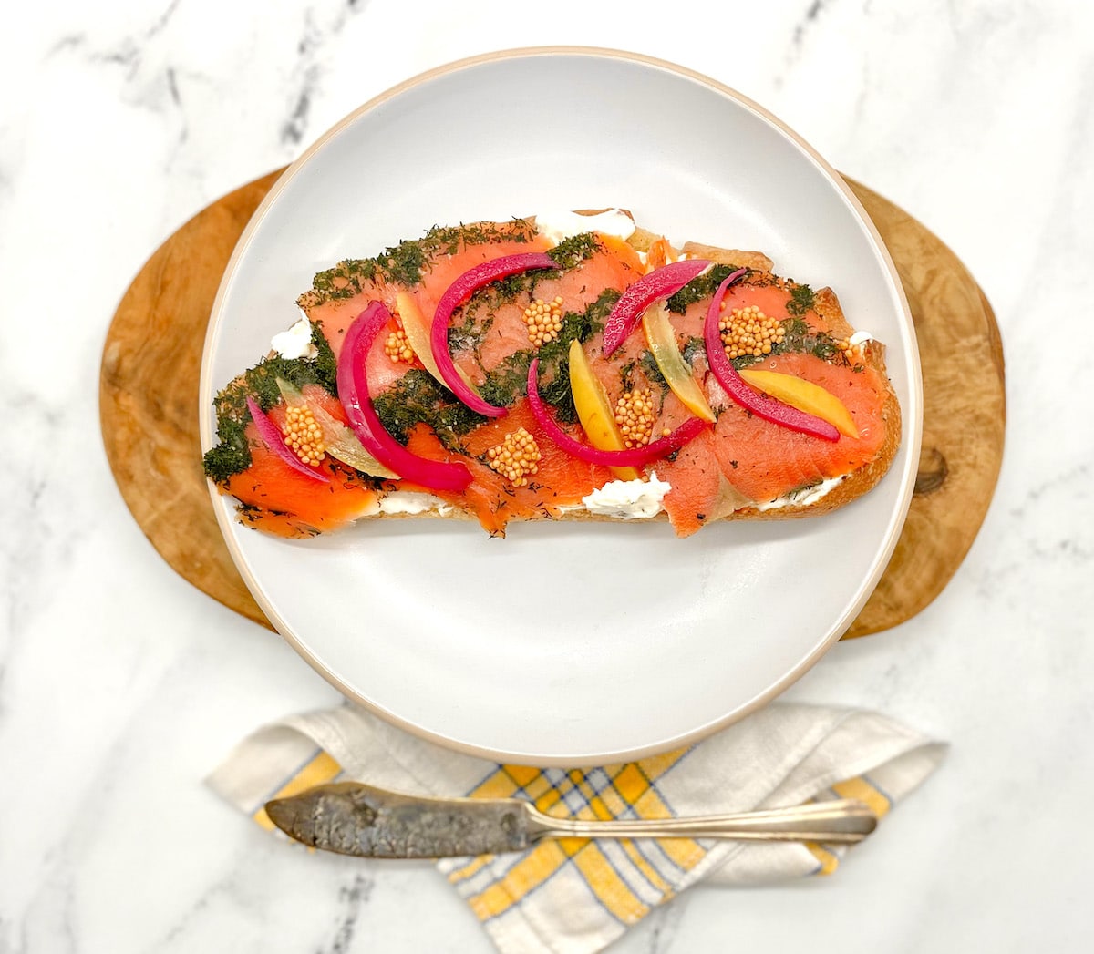 Toasted bread spread with cream cheese and topped with herb-cured salmon, pickled red onion slices, preserved lemon slices and pickled mustard seeds on a white plate with a butcher block and white marble background.