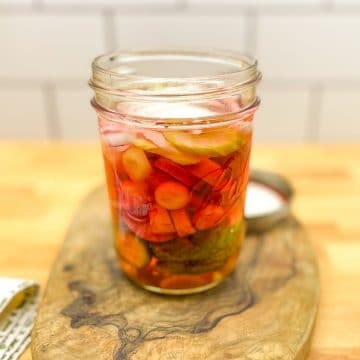 Glass jar filled with pickled onion, celery, rainbow carrots, radish and garlic on a wood cutting board with a white tile background.