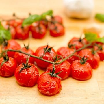 Roasted cherry tomatoes on the vine with roasted garlic cloves and fresh basil leaves on a wooden butcher block cutting board