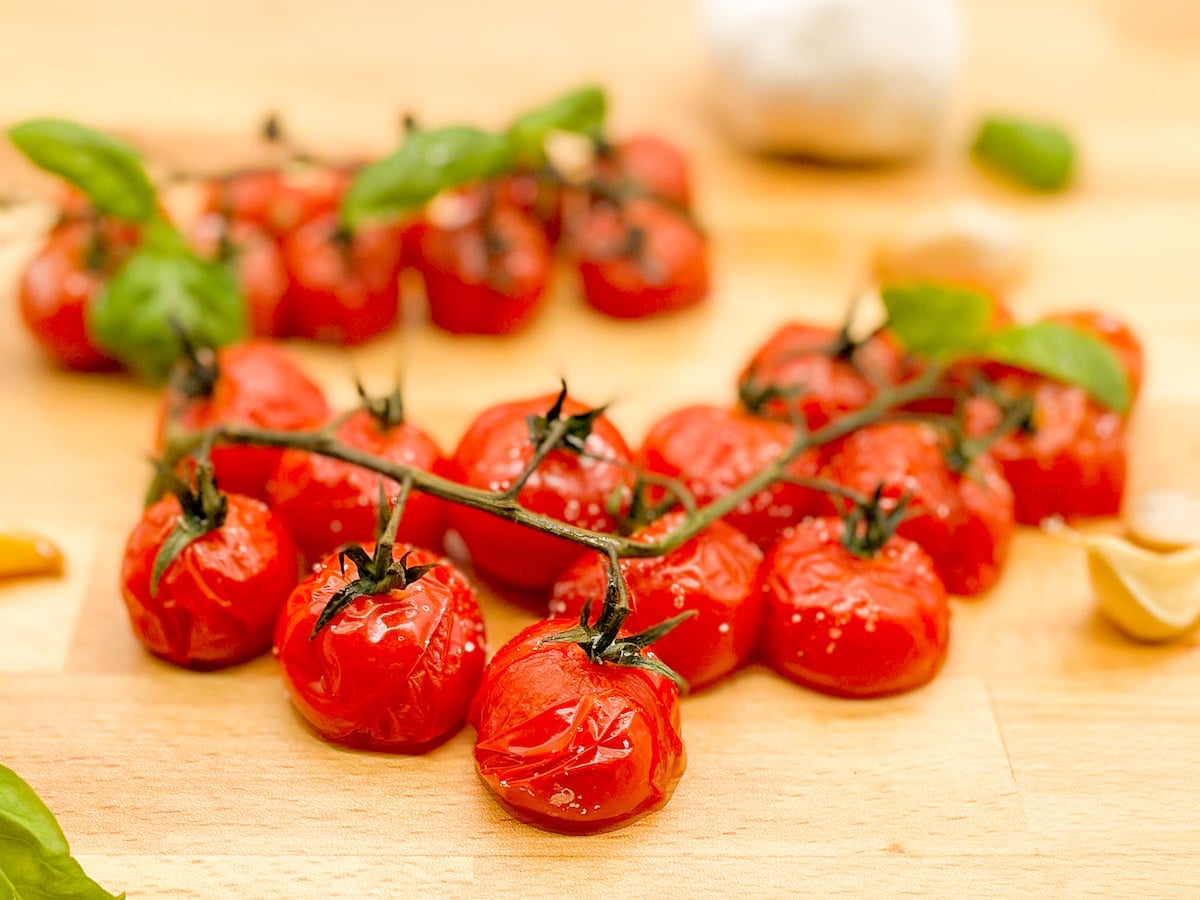 Roasted cherry tomatoes on the vine with roasted garlic cloves and fresh basil leaves on a wooden butcher block cutting board