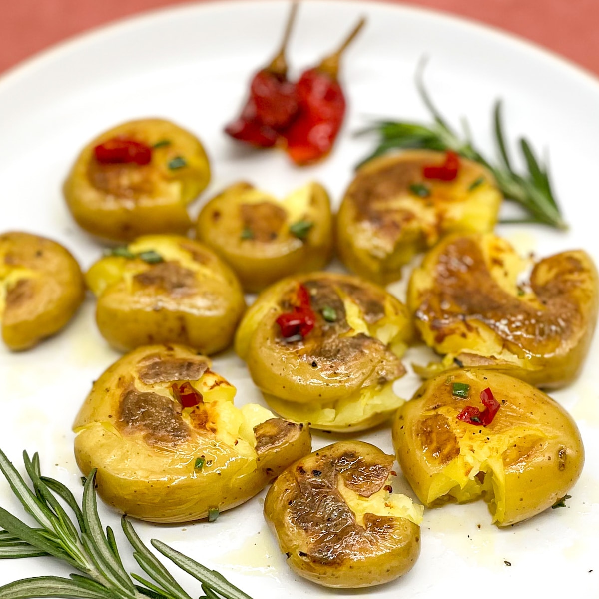 Smashed, pan-fried baby yellow potatoes with rosemary and Calabrian chilis coated in a vinaigrette on white plate with a red background.