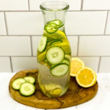 A clear glass pitcher of water with floating slices of lemon, cucumber, ginger and jalapeno on a wood cutting board on a marble countertop