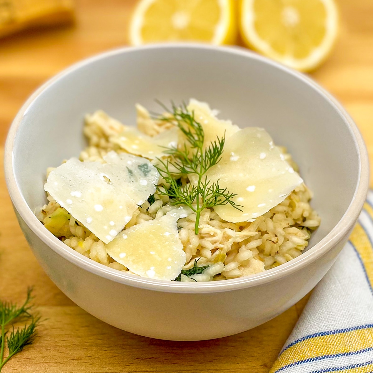 Chicken and leek risotto topped with fresh dill and shaved parmesan in a white bowl on a wood cutting board surrounded by lemons, fresh dill and a yellow and blue gingham napkin.