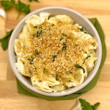 Macaroni and cheese with a toasted bread crumb and parsley topping on a wooden cutting board with a green napkin, parsley and garlic cloves.
