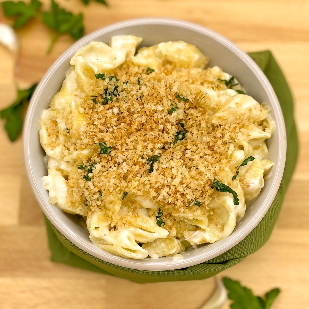 Mac and cheese without milk with a toasted bread crumb and parsley topping on a wooden cutting board with a green napkin, parsley and garlic cloves.