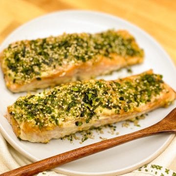 Two baked salmon fillets coated in furikake seasoning on a white plate with a wooden fork and vintage decorate napkin on butcher block.