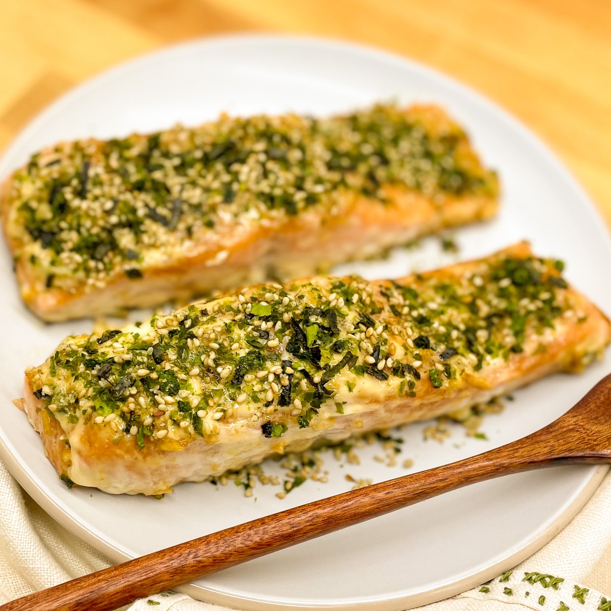 Two baked salmon fillets coated in furikake seasoning on a white plate with a wooden fork and vintage decorative napkin on butcher block.