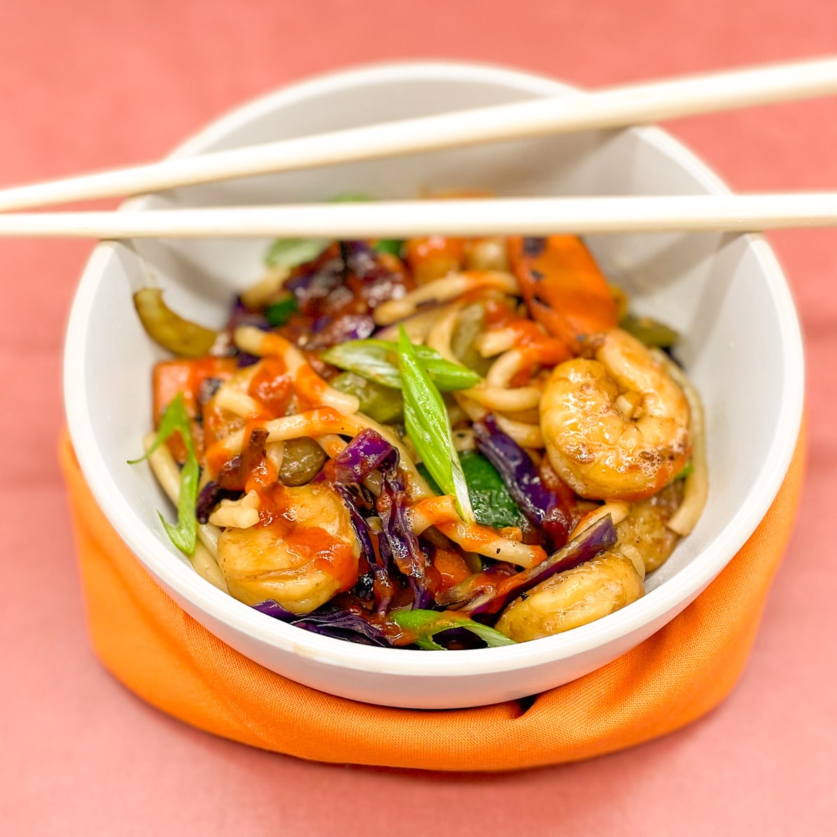 Wok-fried shrimp, udon noodles, carrot, celery, red cabbage and green onion in a white bowl with chopsticks on a red background with an orange napkin.