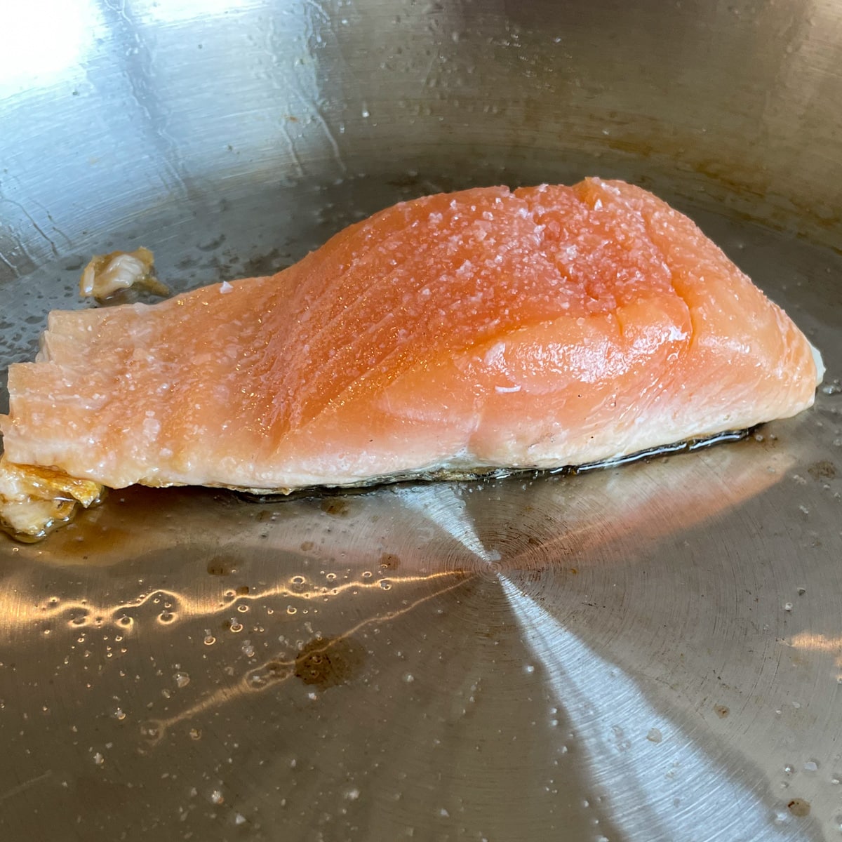 A fillet of salmon is cooking in a stainless steel pan with olive oil and salt. The salmon changes color halfway through, showing it is half cooked.