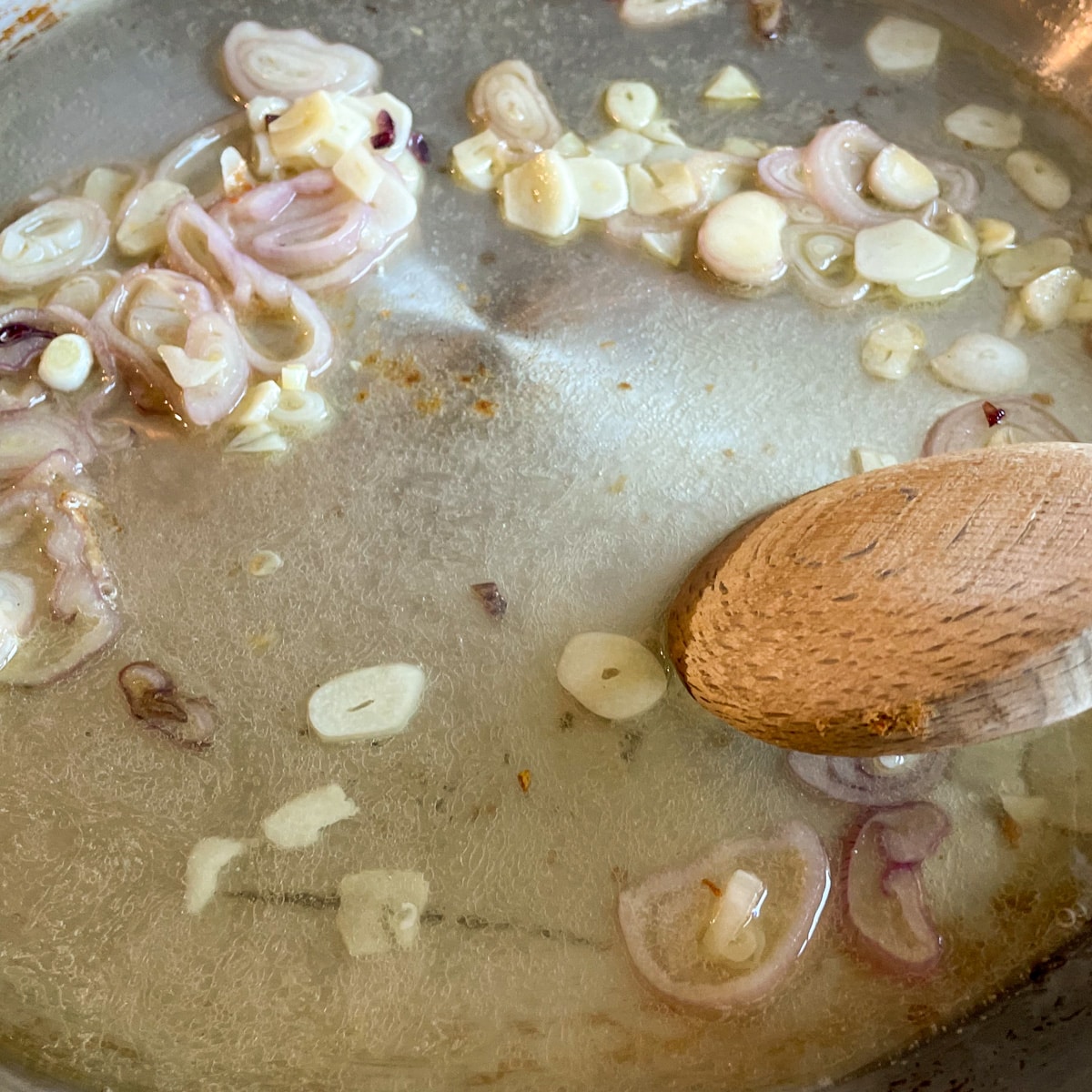 Sautéed shallots and garlic are shown in a stainless steel pan with white wine and lemon juice with a wooden spoon mixing the ingredients together.
