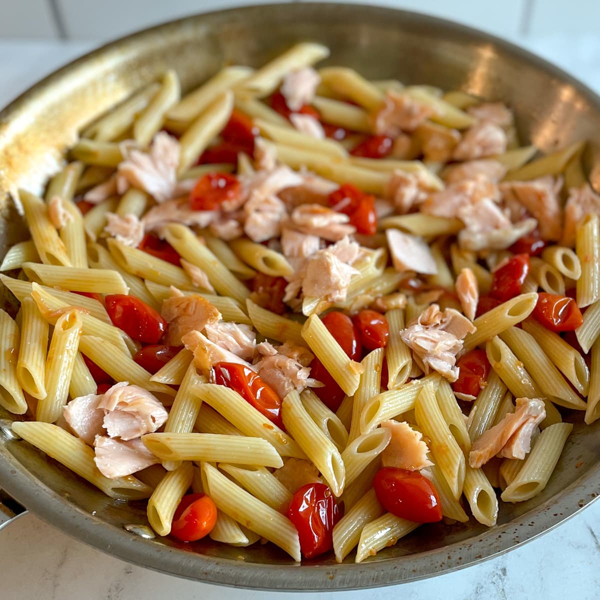 Penne pasta mixed with flaked salmon and burst cherry tomatoes in a stainless steel pan over a white marble countertop.