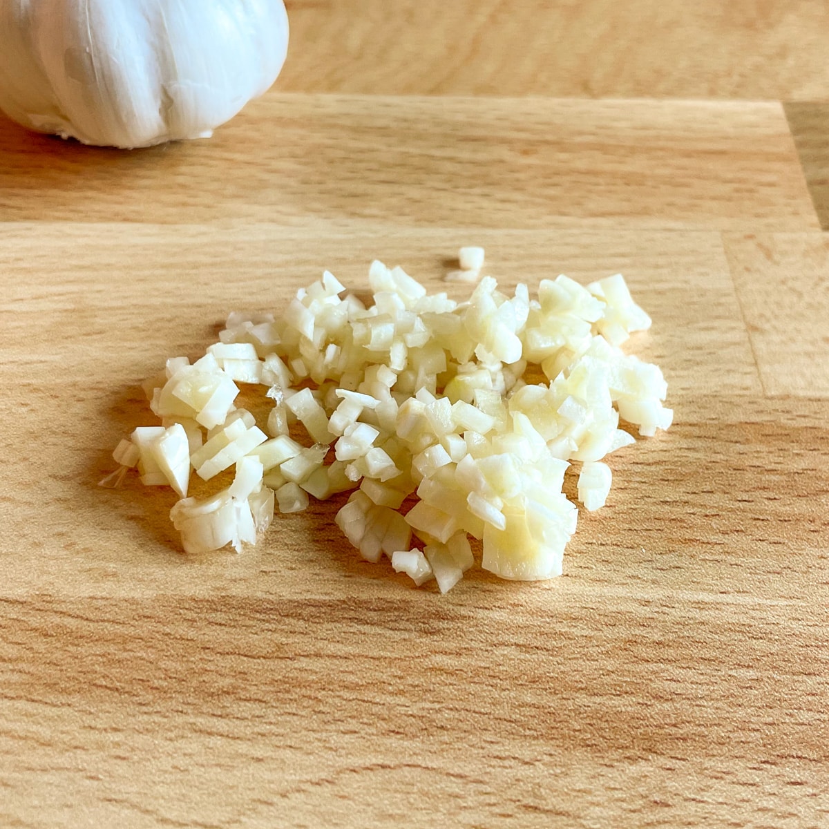 Minced garlic with a head of garlic in the background on a butcher block cutting board