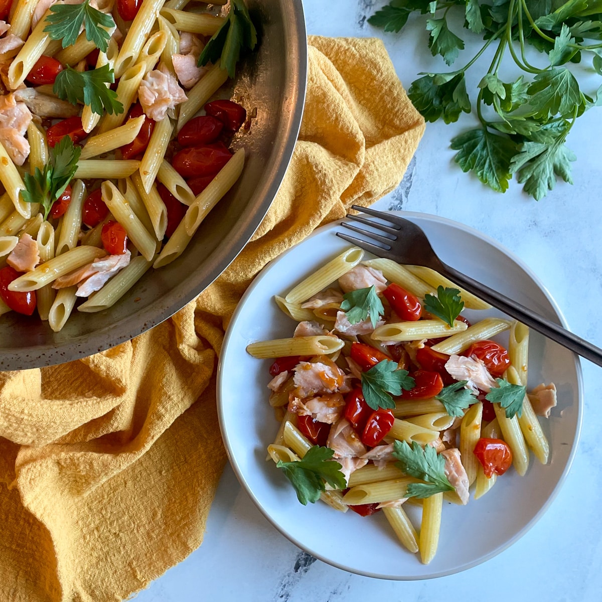 A white plate of penne pasta with burst cherry tomatoes and flaked salmon topped with parsley sits on a white marble countertop with a yellow linen, a pan of salmon penne pasta, a bunch of parsley, and a copper-colored fork.
