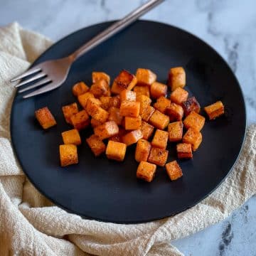 Cubed, sautéed sweet potatoes sit on a black plate with a gold fork on a white marble countertop with an off white linen.