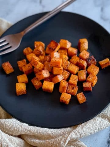 Cubed, sautéed sweet potatoes sit on a black plate with a gold fork on a white marble countertop with an off white linen.