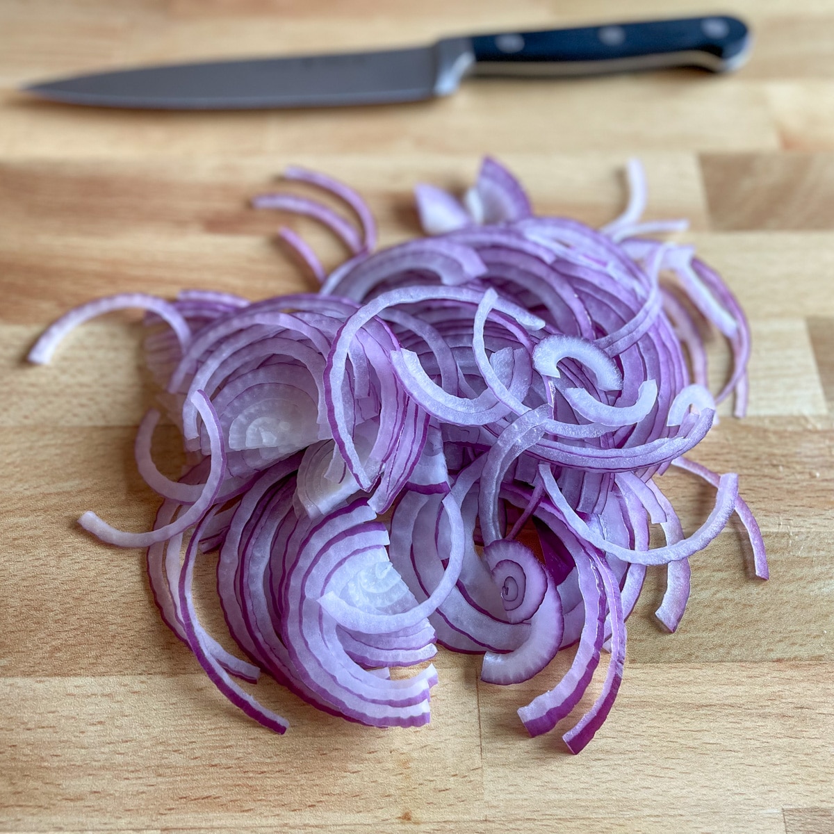 Sliced red onions sit on a wooden cutting board with a kitchen knife in the background.
