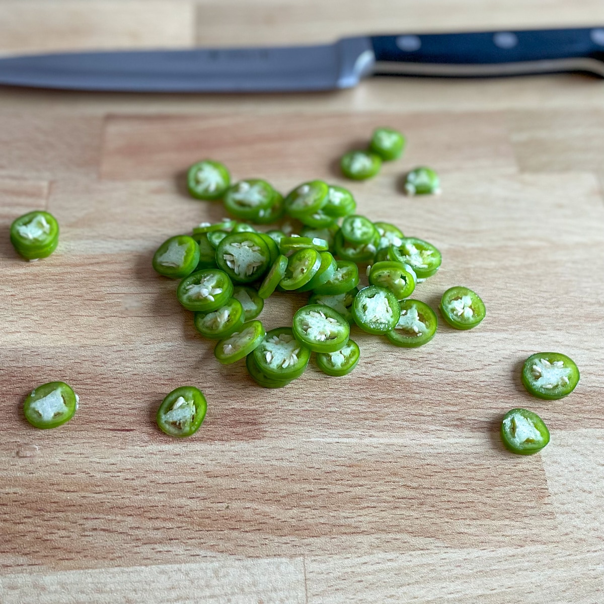 Sliced serrano peppers sit on a wooden cutting board with a kitchen knife in the background.