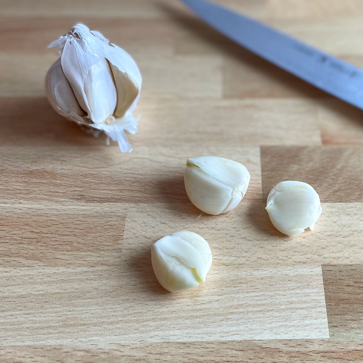 Three cloves of smashed garlic sit in front of a partial head of garlic and a kitchen knife on a wooden cutting board.