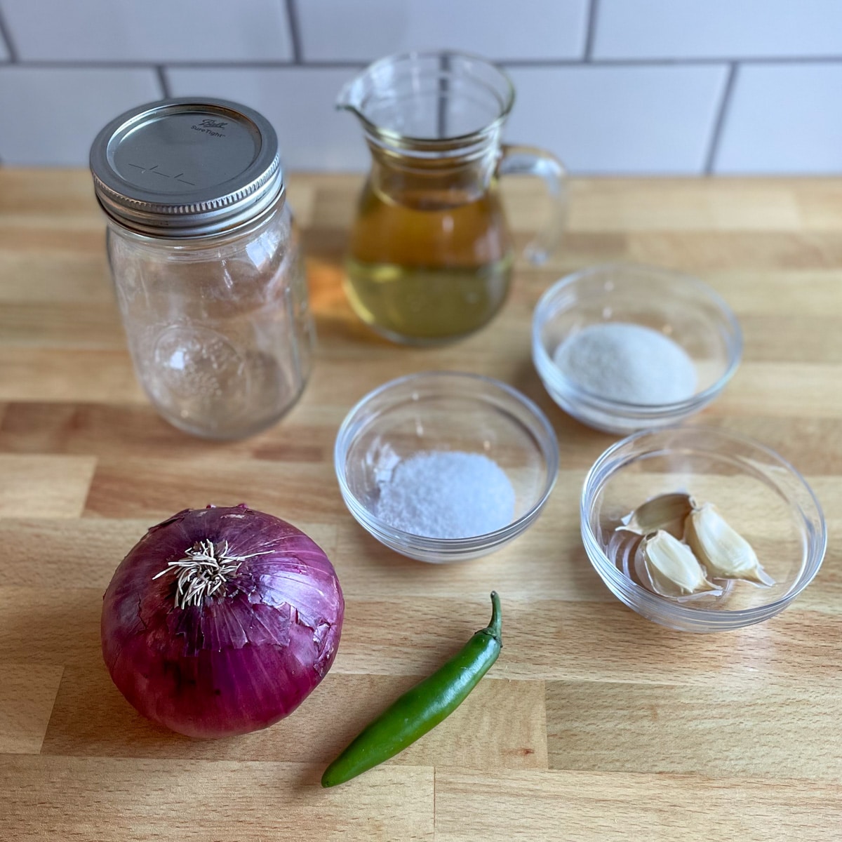 Red onion, serrano peppers, small Mason jar, small clear pitcher of rice vinegar, and three clear pinch bowls of salt, sugar, and garlic cloves sit on a wooden cutting board.