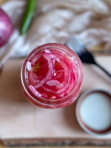 Sliced red spicy pickled onions are shown in a glass jar on a rustic wood cutting board with a lid, a black fork, a white linen, a red onion, and a Serrano pepper in the background.