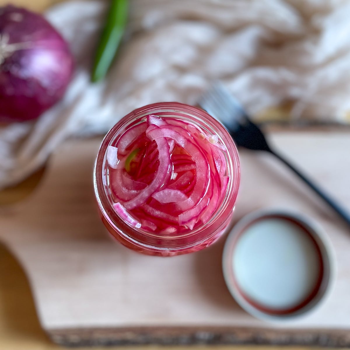 Sliced red spicy pickled onions are shown in a glass jar on a rustic wood cutting board with a lid, a black fork, a white linen, a red onion, and a Serrano pepper in the background.