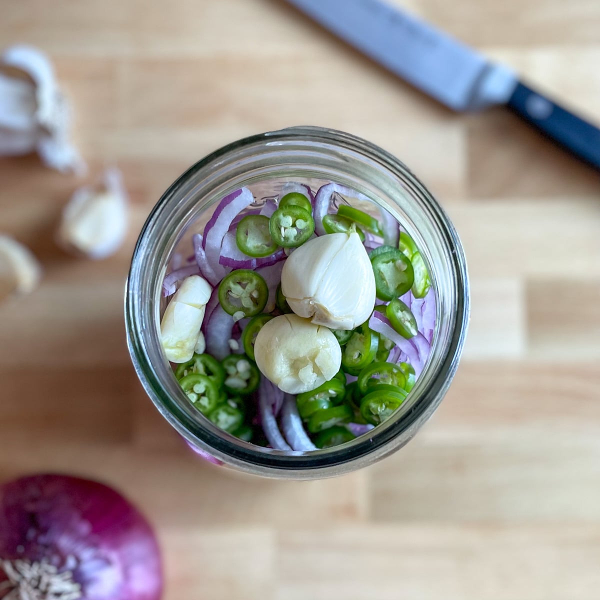 A clear glass jar with sliced red onion, sliced Serrano peppers, and three cloves of smashed garlic sit on a wooden cutting board surrounded by cloves of garlic, a red onion, and a kitchen knife.