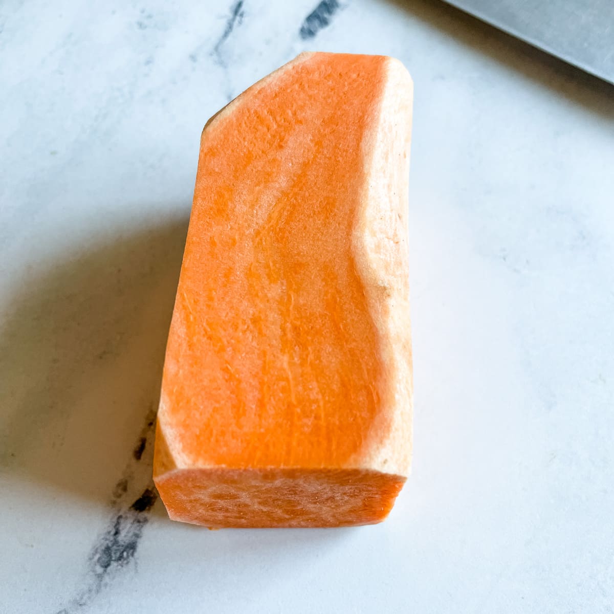 A peeled sweet potato with the round sides trimmed to create a rectangle sits on a white marble counter top next to the blade of a kitchen knife.