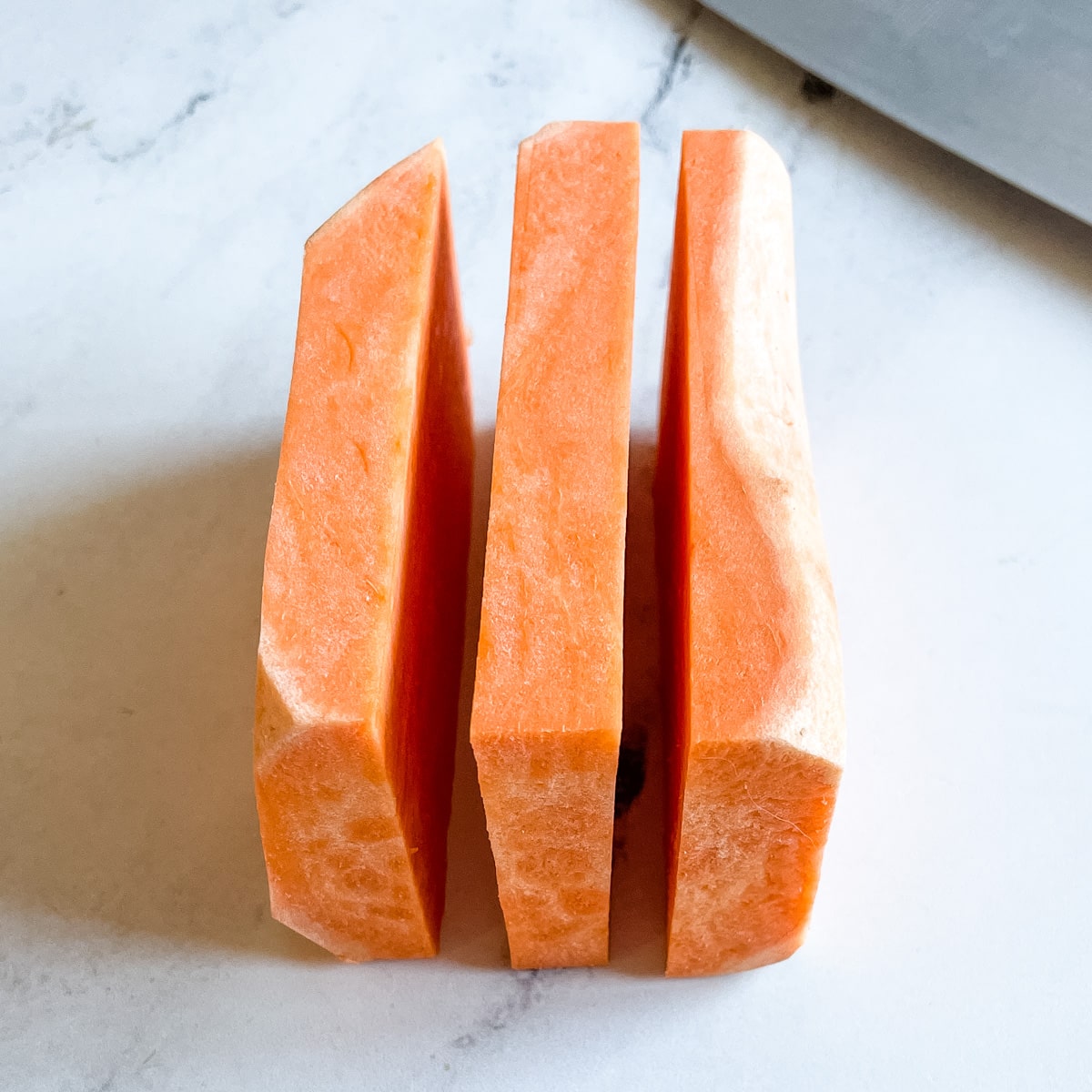 A peeled sweet potato cut horizontally into 3 equal pieces sits on a white marble counter top next to the blade of a kitchen knife