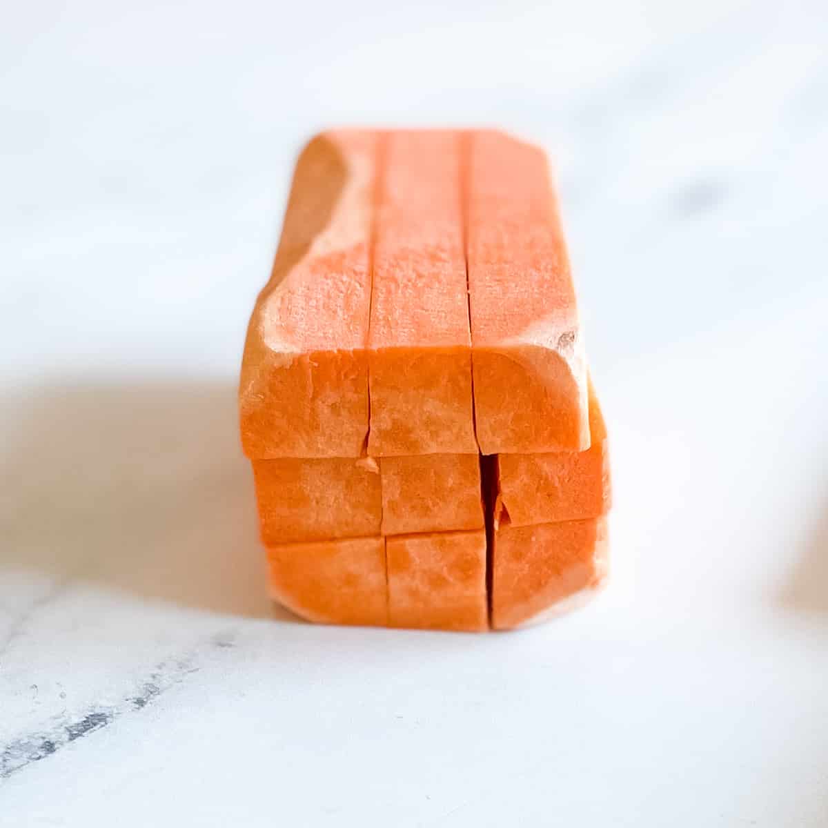 A peeled sweet potato cut both horizontally and diagonally into equal pieces to create long sticks sits on a white marble counter top.