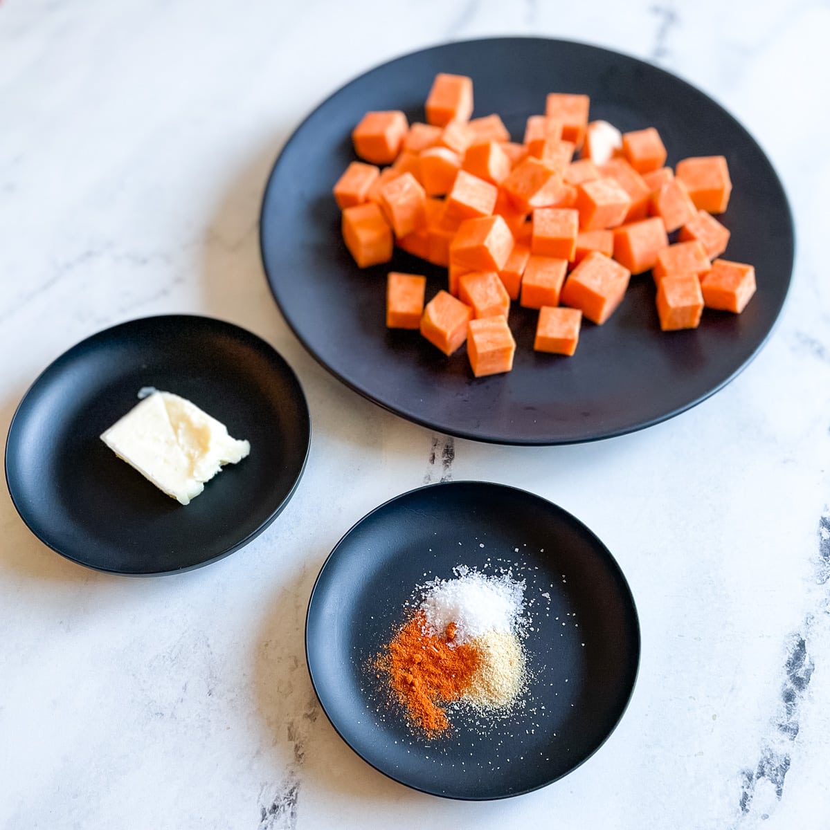 A large black plate with cubed sweet potatoes, a small black plate with a pat of butter, and a small black plate with cayenne, garlic powder, and salt sit on a white marble counter top
