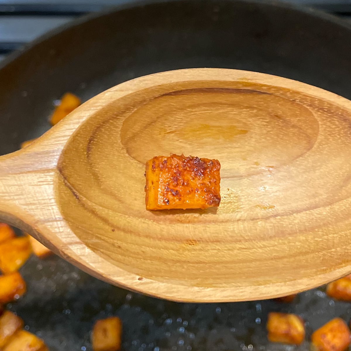 A wooden spoon in the foreground shows a browned cube of sweet potato over a black frying pan with sweet potato cubes cooking in it.