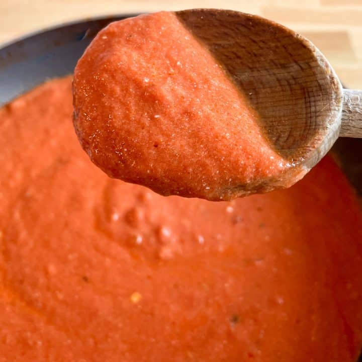 A wooden spoon of tomato and mascarpone sauce is held in the foreground over a pot of cooking sauce in a black pan.