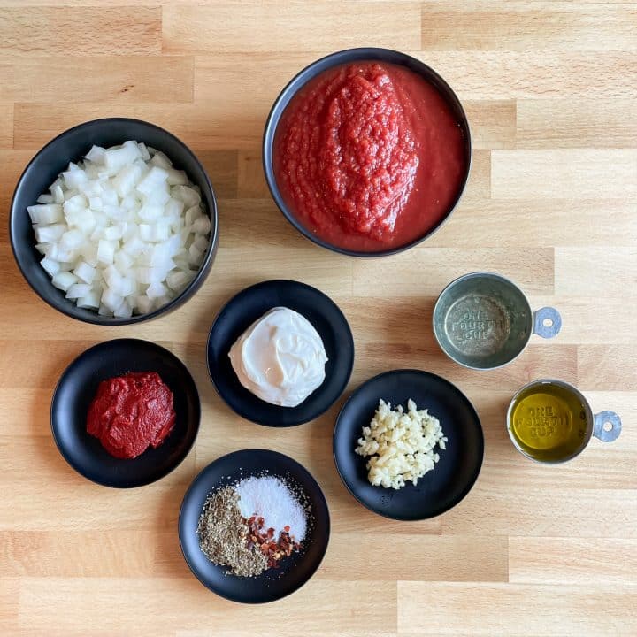 Two black bowls hold crushed tomatoes and diced onion; four small black plates hold mascarpone, tomato paste, minced garlic, and a spice mixture of Italian seasoning, salt, and red pepper flake; two small metal measuring cups hold white wine and olive oil. All ingredients are on a butcher block cutting board.