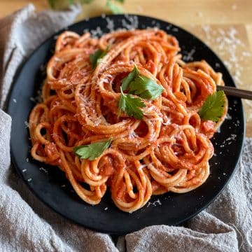 Twirled spaghetti on a black plate topped with parsley and parmesan cheese on a butcher block cutting board with a light gray linen