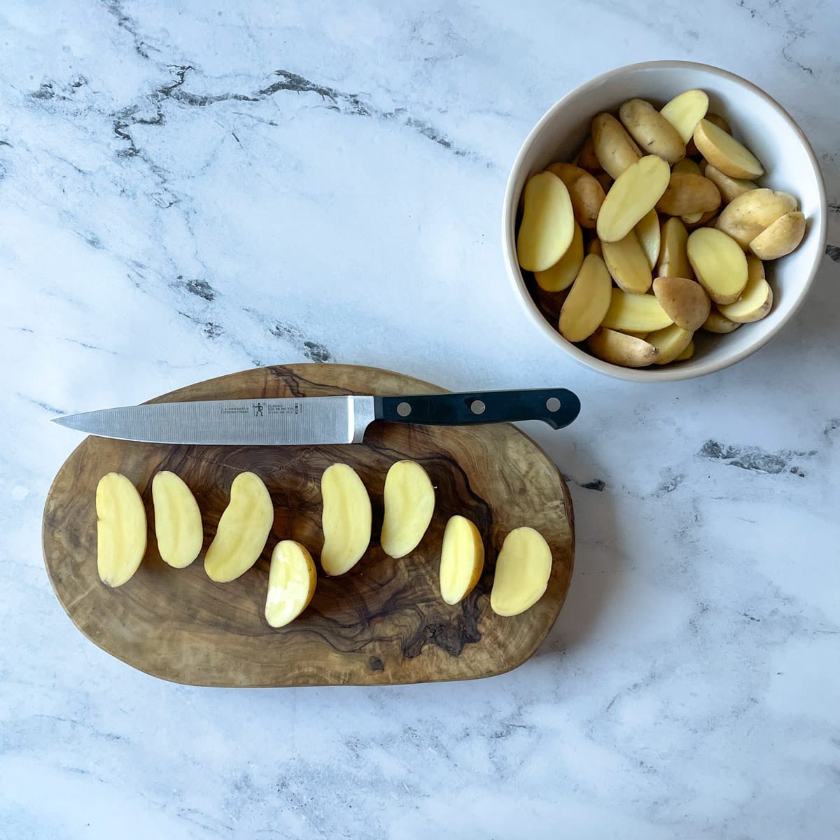 Fingerling potatoes cut lengthwise on a wood cutting board with a bowl of cut potatoes in the background on a white marble counter.