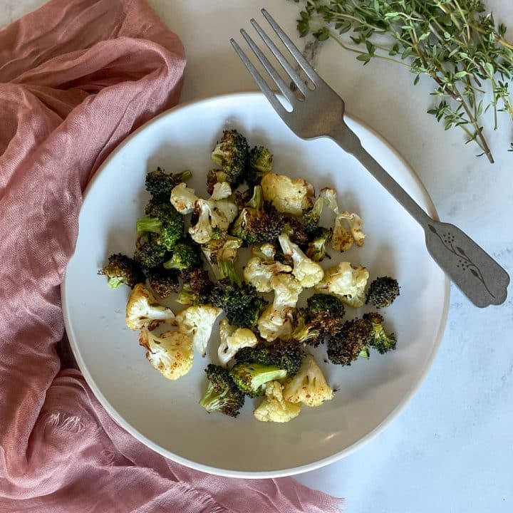 Roasted broccoli and cauliflower sit on a white plate with a decorative silver fork and a dusty pink linen on a white marble counter with a bunch of fresh thyme.