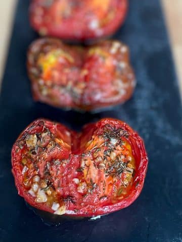 Three red and purple heirloom roasted air fryer tomatoes sit on a black slate cooking stone on a butcher block cutting board.