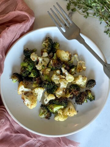 Roasted broccoli and cauliflower sit on a white plate with a decorative silver fork and a dusty pink linen on a white marble counter with a bunch of fresh thyme.
