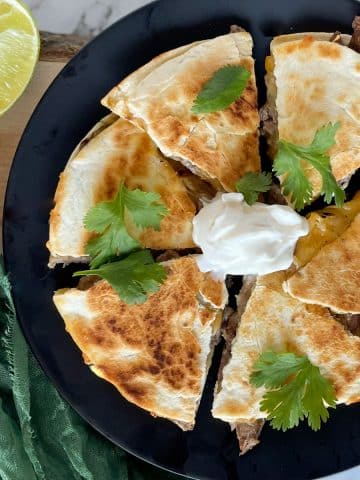 Carne asada quesadilla cut into six pieces and topped with cilantro and cream on a black plate surrounded by a green linen, a lime slice, and wooden cutting board.