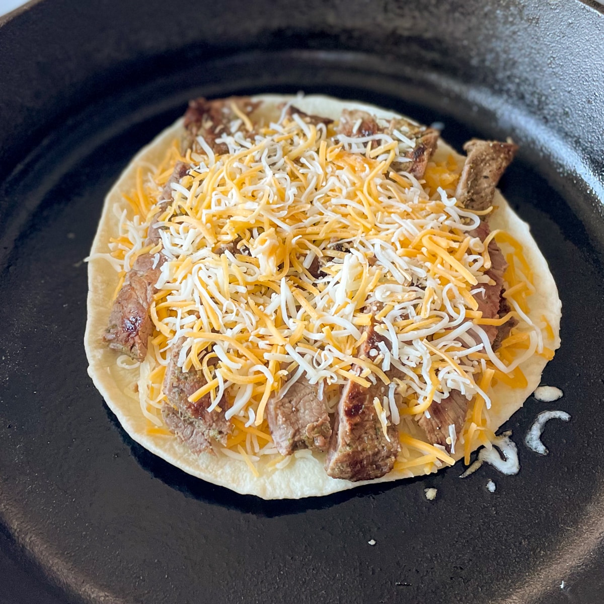 A tortilla, shredded cheese, and sliced carne asada are stacked in a cast iron pan.