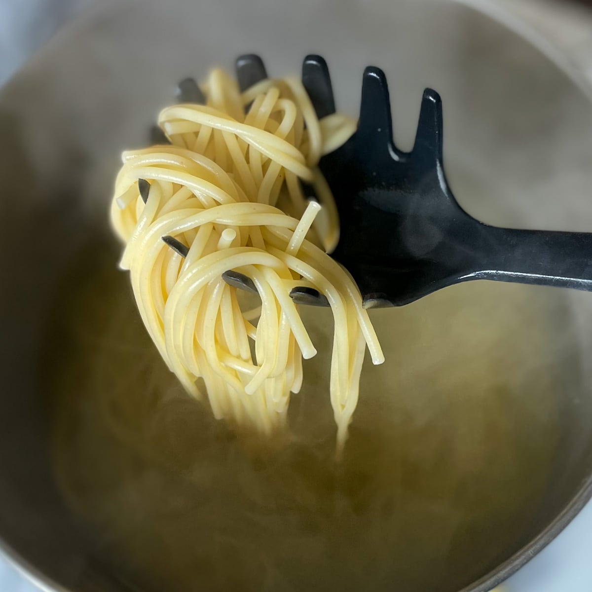 Cooked spaghetti is held on a black spaghetti serving spoon above a silver pot of boiling water.