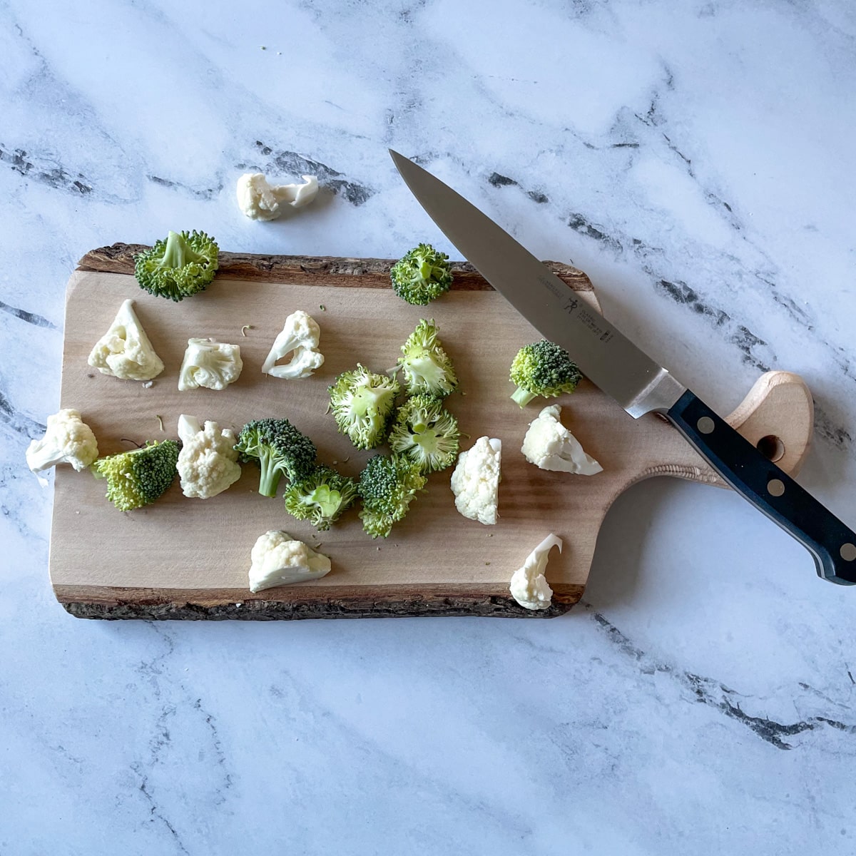 Cut broccoli and cauliflower cut into one inch florets sit on a wooden cutting board with a kitchen knife on a marble counter.