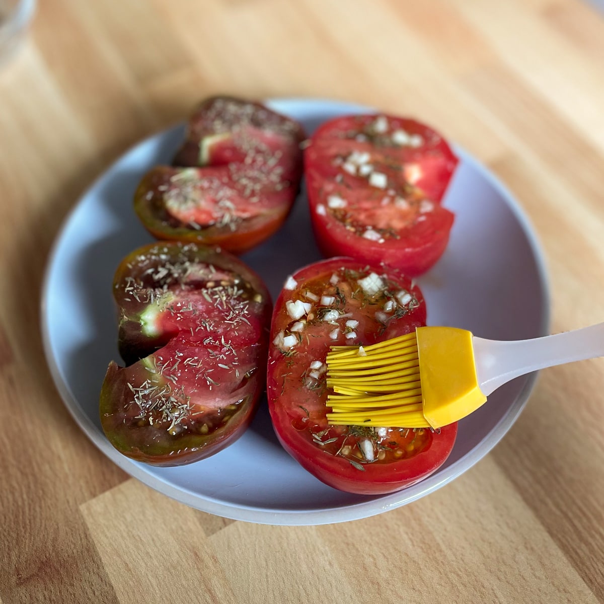 Minced garlic is spread on the cut side of a halved red heirloom tomato with a yellow silicone brush.