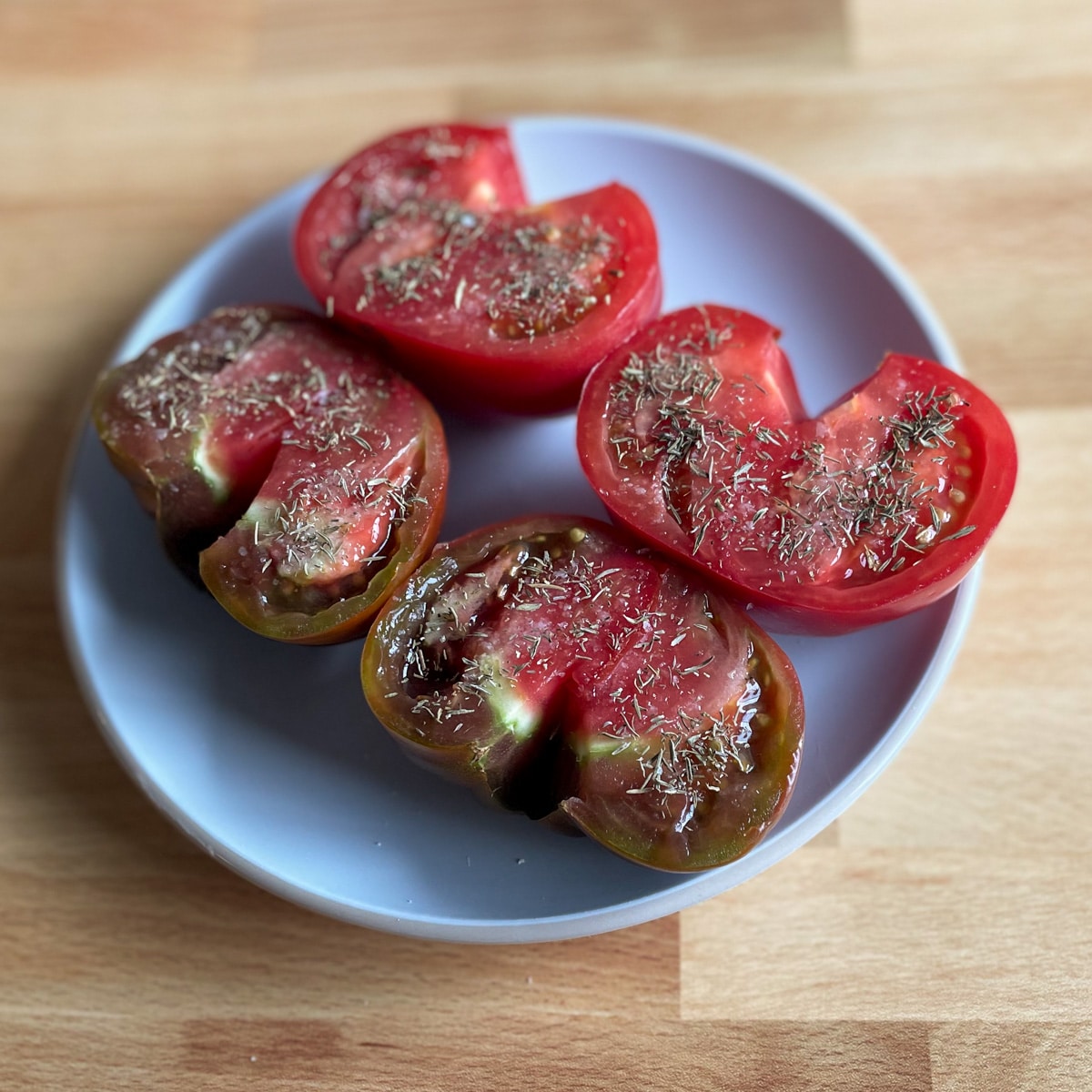 One halved red heirloom tomato and one halved purple and red heirloom tomato dusted with salt and dried thyme sit on a white plate on a butcher block cutting board.