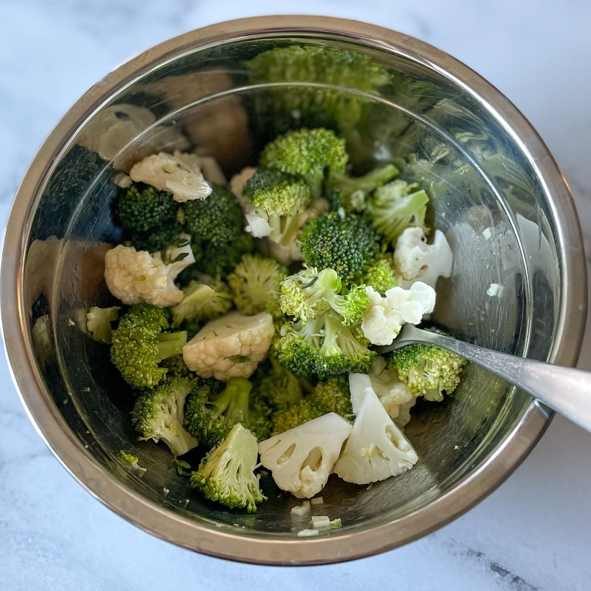 Broccoli and cauliflower coated in lemon garlic thyme vinaigrette are shown in a metal bowl with a fork on a marble counter top.