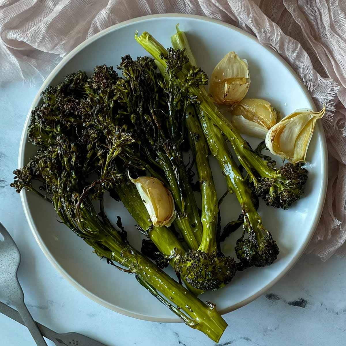 Roasted broccolini and garlic cloves are shown on a white plate with a light pink linen with two forks.
