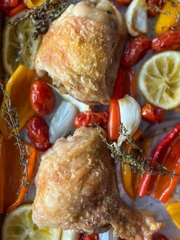 Two crispy skinned chicken thighs sit on a sheet tray surrounded by roasted baby sweet peppers, sliced lemon, quartered shallots, garlic, thyme, and cherry tomatoes.